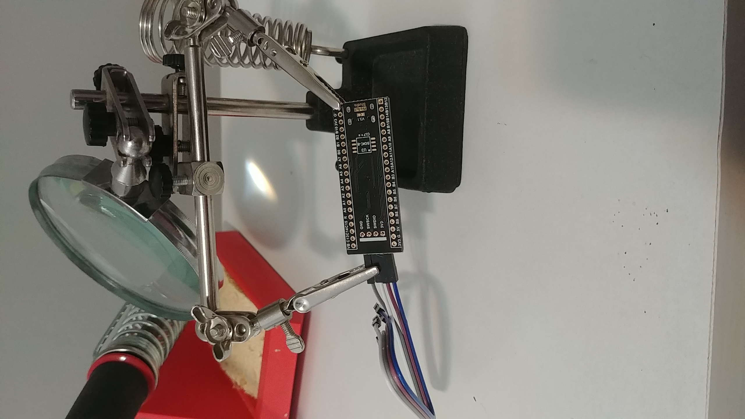 black pill PCB attached to a third hand tool to keep it steady while I solder. The SWD header is put into the board, and has 4 wires connected to it